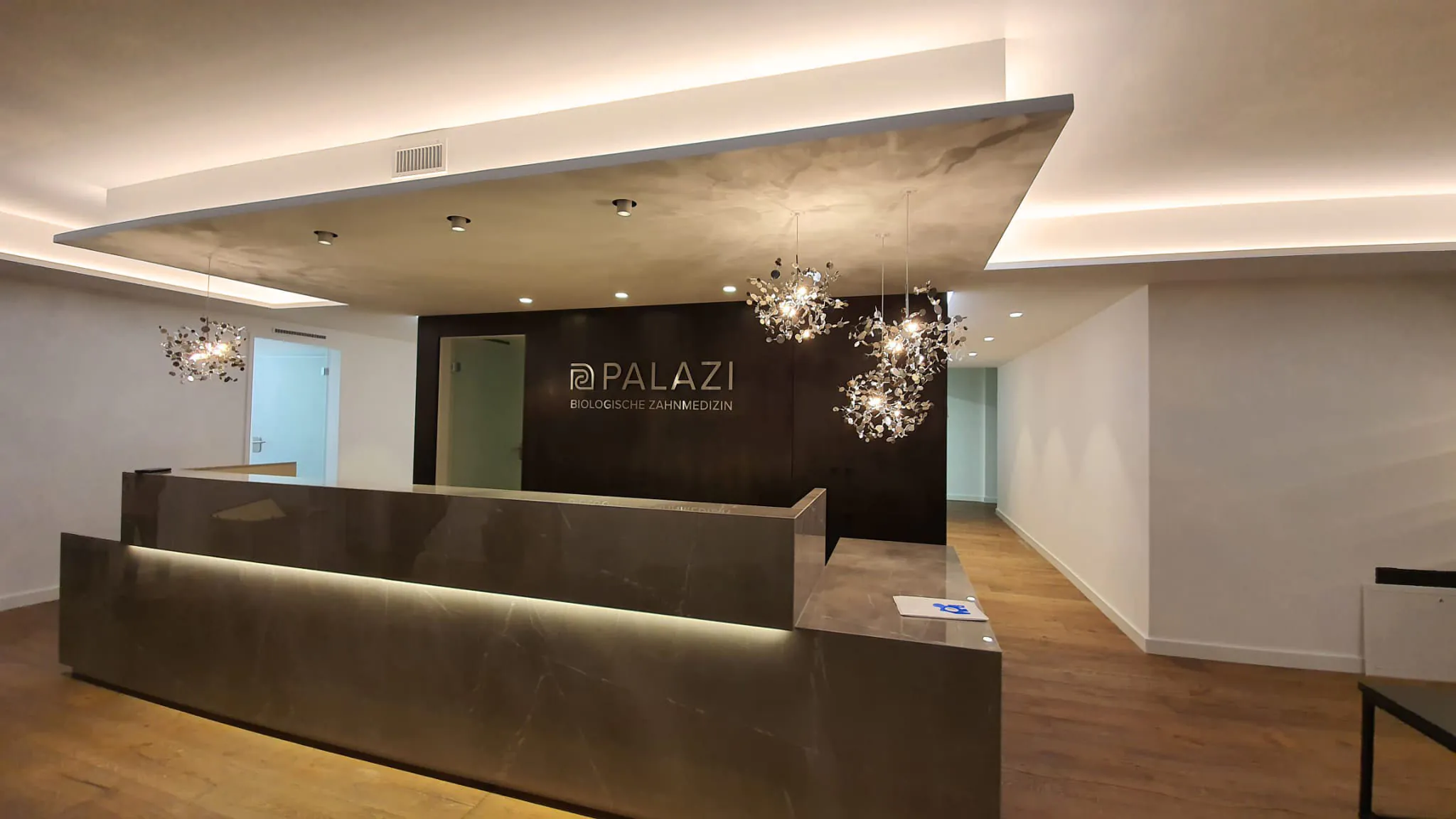 Empfang Theke einer Zahnarztpraxis mit 3 Silberfarbenen Pendelleuchten von Terzani und Einbaustrahlern von Delta Light in der Decke
