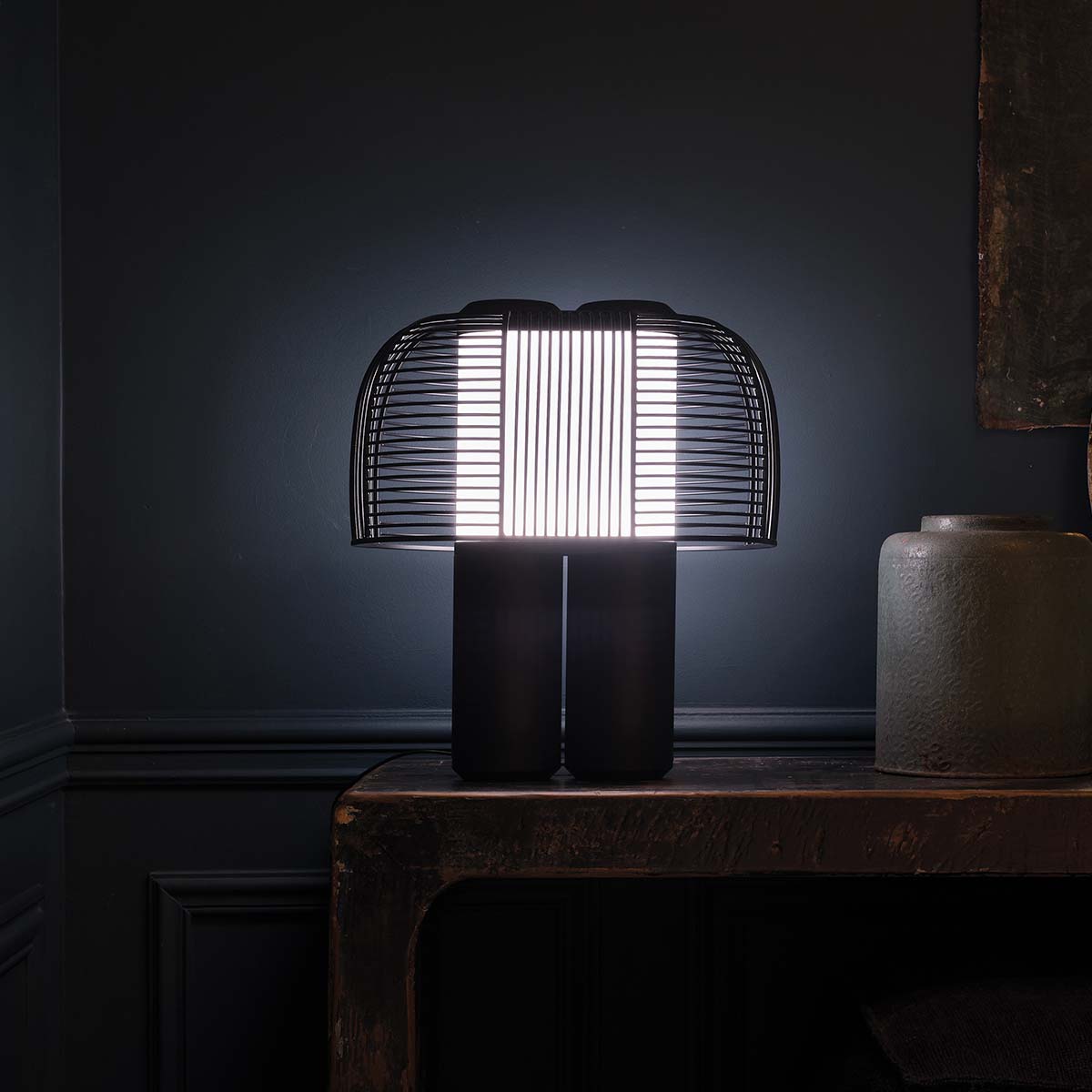 Schwarze Yasuke-Tischleuchte leuchtet auf einem dunklen Holztisch vor einer dunkelblauen Wand.