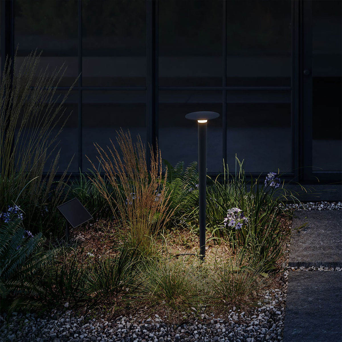 Längliche Erdspießleuchte schwarz mit Solarpanel und schwarzem Verbindungskabel im Garten zwischen Pflanzen