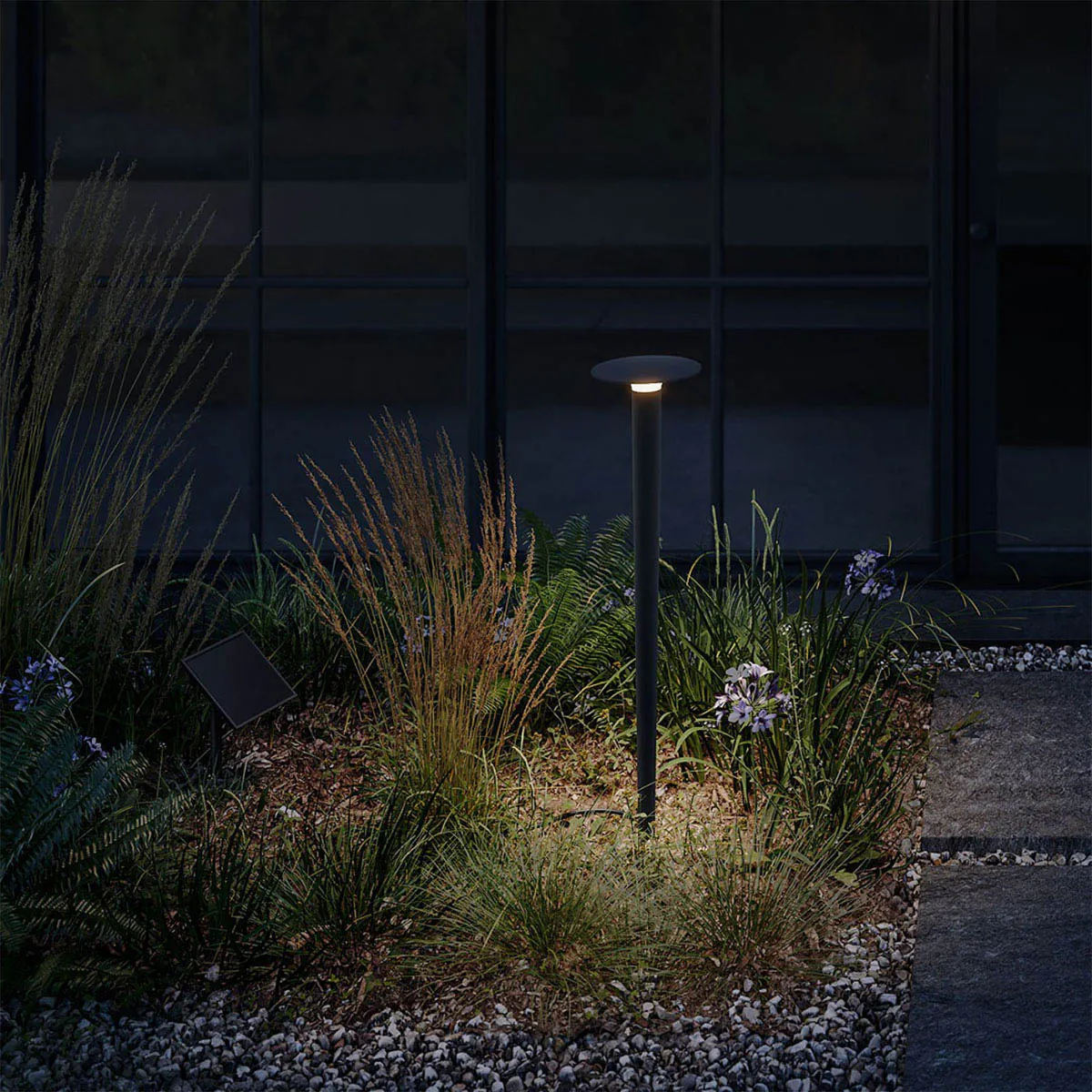 Längliche Erdspießleuchte schwarz mit Solarpanel und schwarzem Verbindungskabel im Garten zwischen Pflanzen