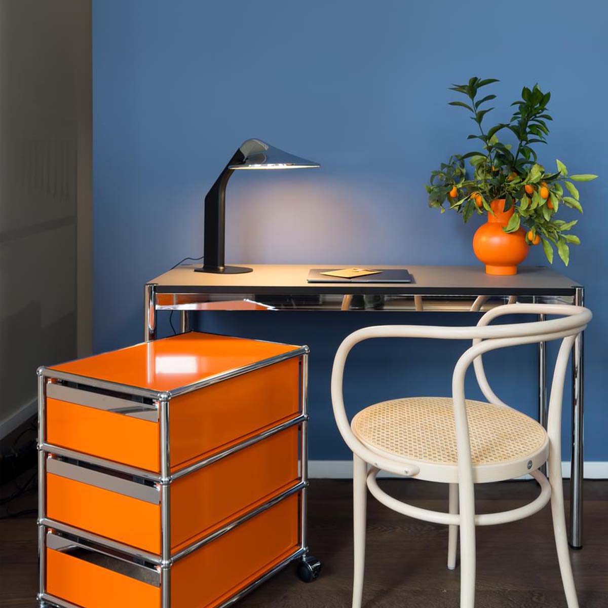Niwaki-Tischleuchte auf einem Schreibtisch vor blauer Wand, daneben ein weißer Stuhl und eine orange Kommode.