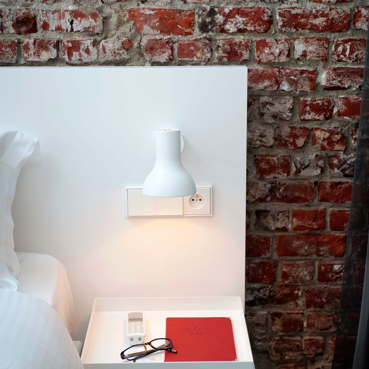 Anglepoise Type 75 Mini Wandleuchte, Ø: 13 cm, H: 15 cm im Wohnambiente: weiße Mini-Wandleuchte über Nachttisch vor Backsteinwand mit warmem Licht und minimalistischem Schlafzimmerstil.