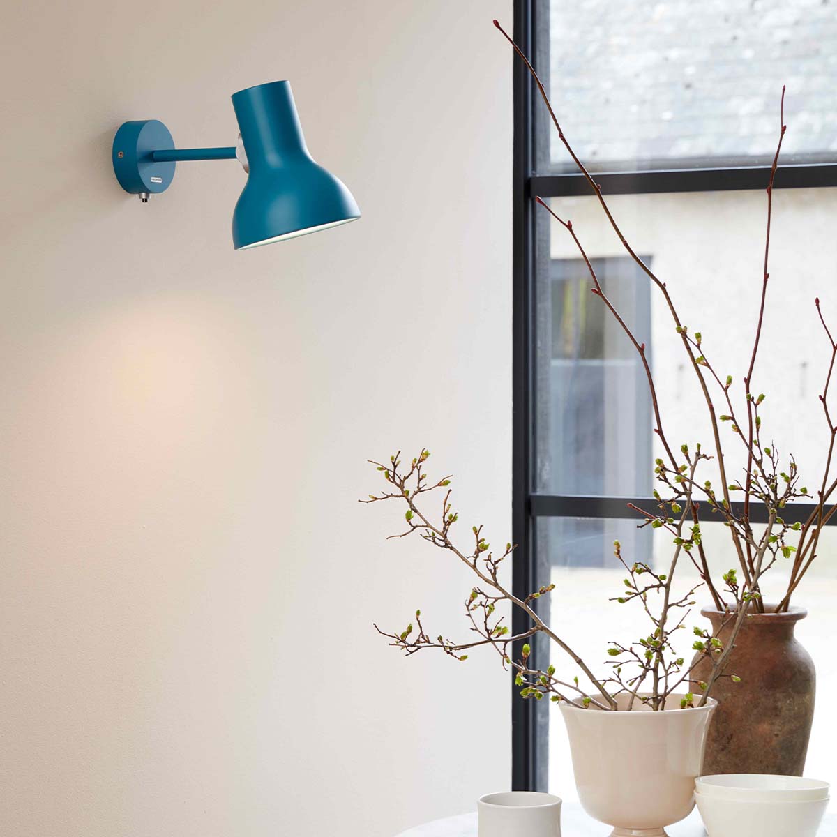 Anglepoise Type 75 Mini Wandleuchte Margaret Howell Edition im Wohnambiente: türkisblaue Wandleuchte neben großem Fenster mit dekorativen Zweigen und modernem Wohnambiente.